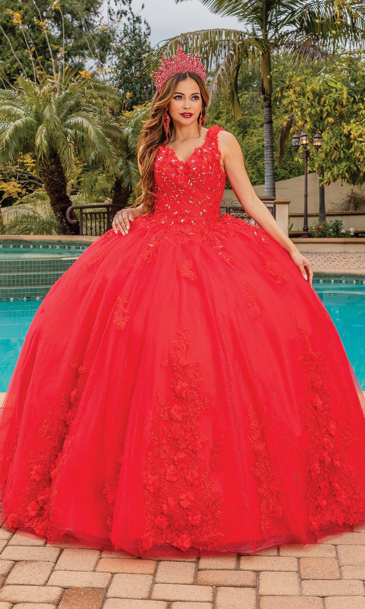 A woman poses by a pool in the vibrant Quinceanera Dress 1623 By Dancing Queen, featuring a bright red floral design, voluminous skirt, and matching crown amidst palm trees and lush greenery.